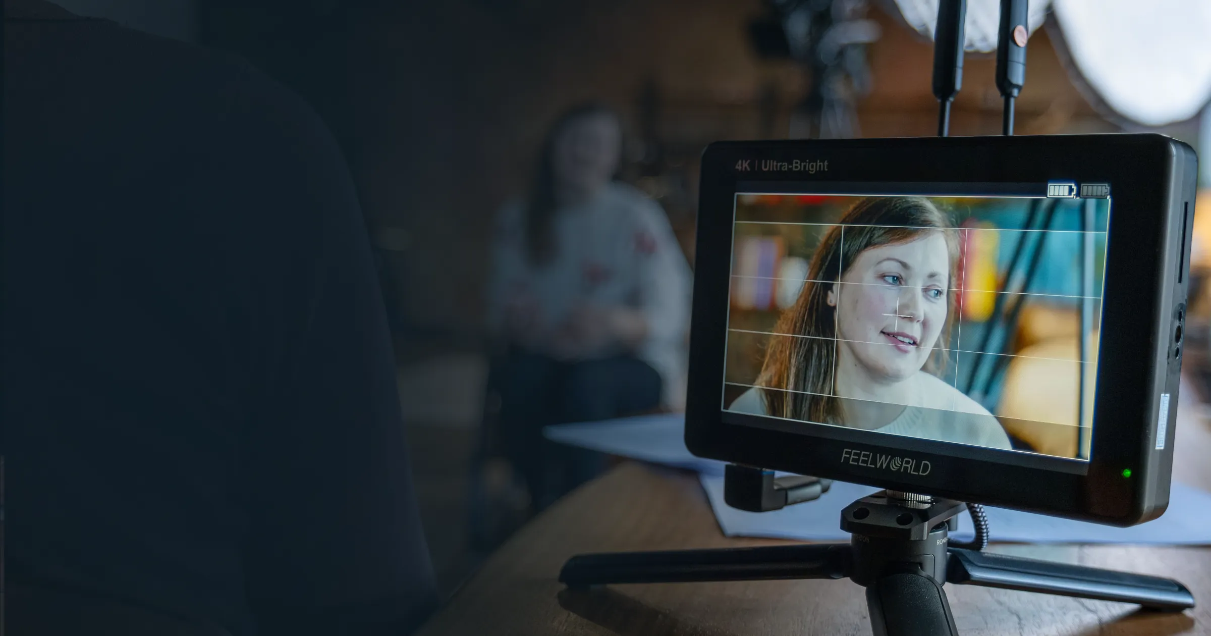 Close-up of a Feelworld 4K Ultra-Bright camera monitor on a tripod showing a young woman being filmed for the Cafe Lingo Oksana's Story video production, with studio lighting and blurred background